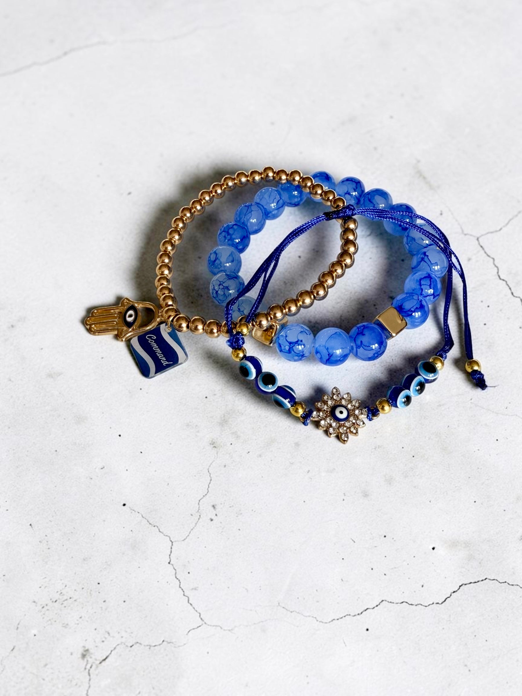Three beaded stretch bracelets in shades of blue and gold, featuring evil eye charms, a Hamsa hand, and a starburst charm, displayed on a light grey textured surface.