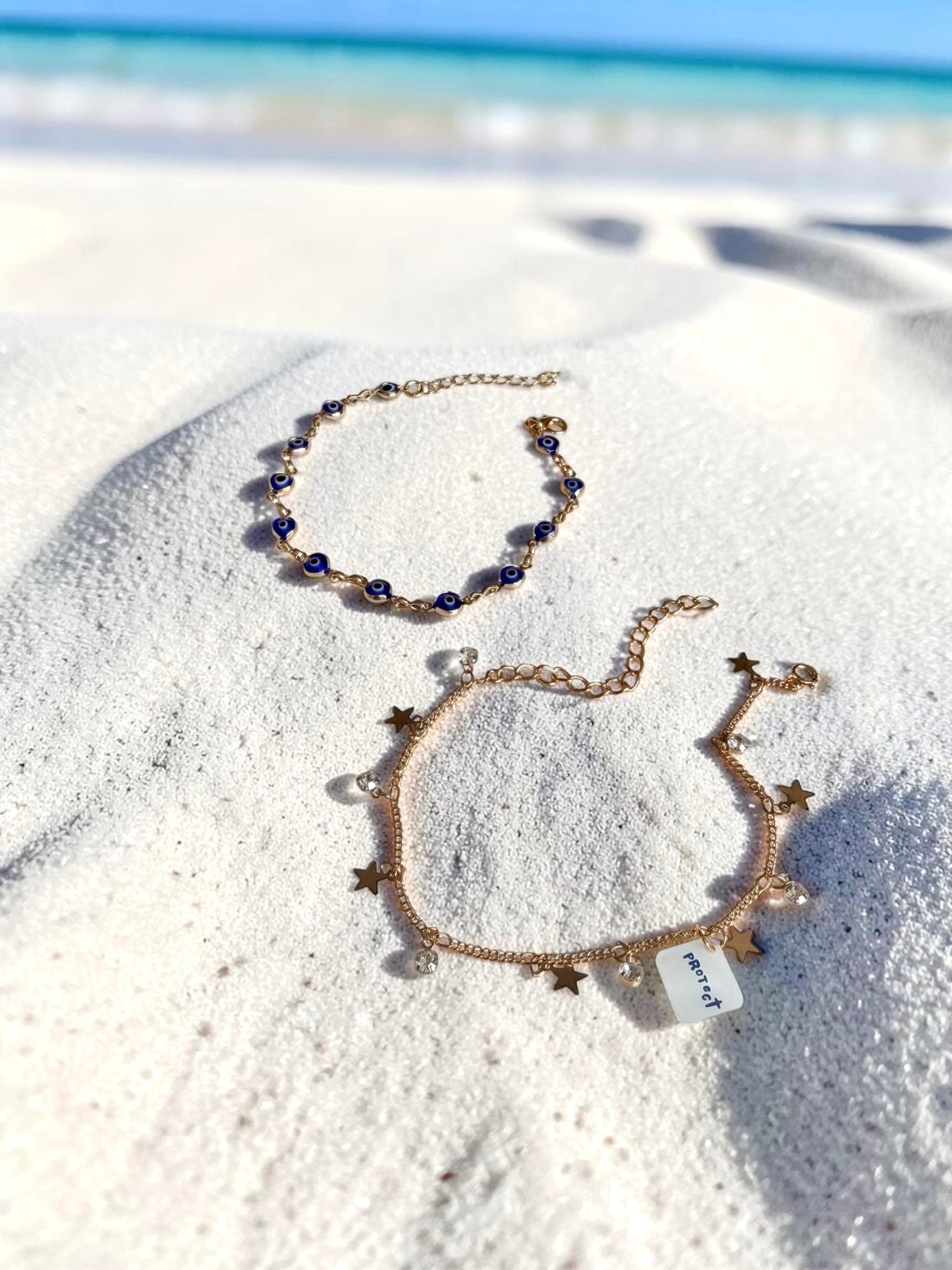 Two delicate gold-toned bracelets displayed on white sand near the ocean. One bracelet features small dark blue evil eye beads, and the other is decorated with gold star charms, cubic zirconia stones and a small white tag labelled 'Protect'.