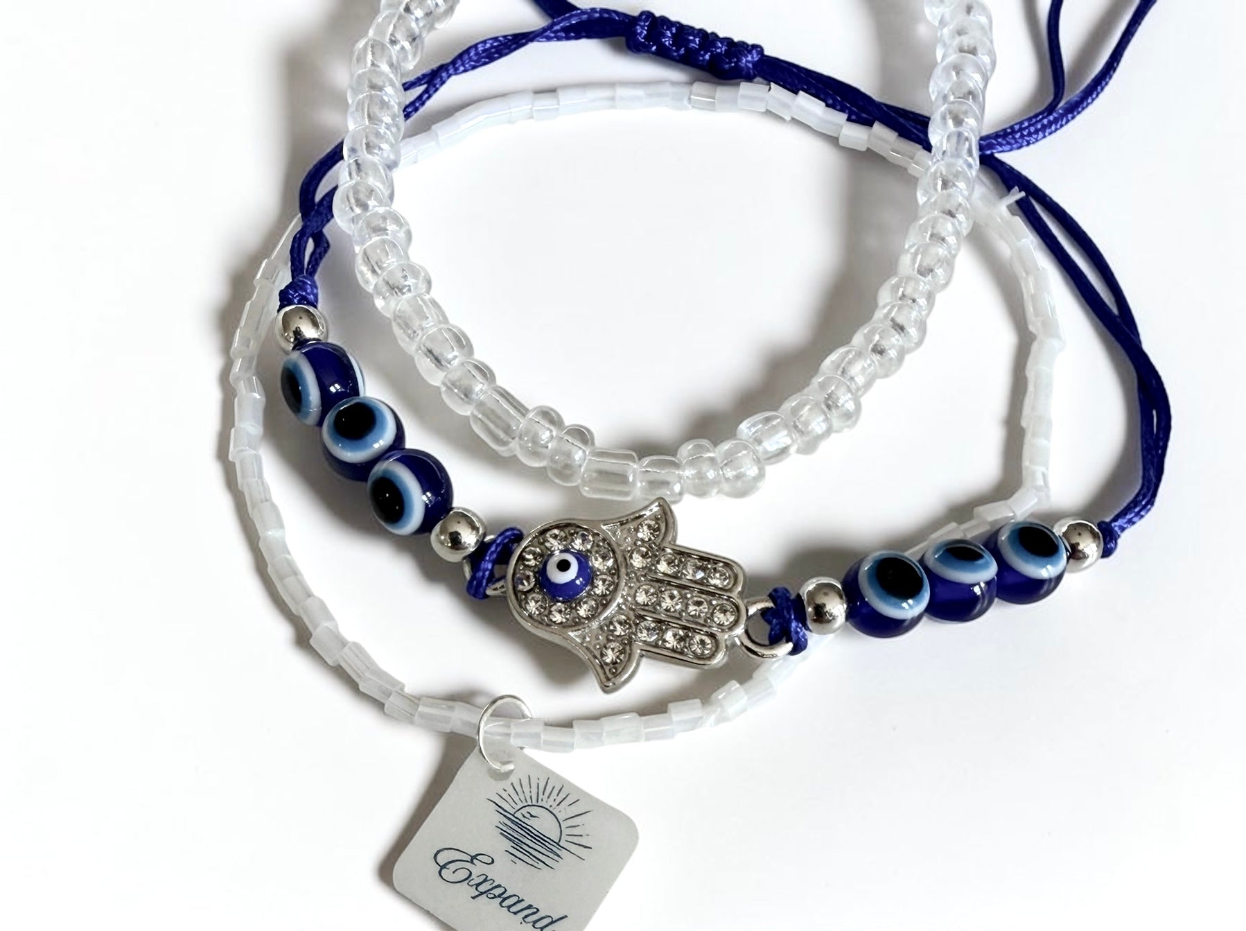A set of three beaded bracelets on a white background. One bracelet is made of clear faceted beads, another is made of dark blue cords with blue and white evil eye beads and a silver Hamsa hand charm, and the third is made of white faceted beads with a tag charm labelled 'Expand'.