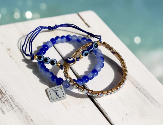 Three blue and gold beaded bracelets with evil eye details, displayed on a white wooden surface with a blurred turquoise background.