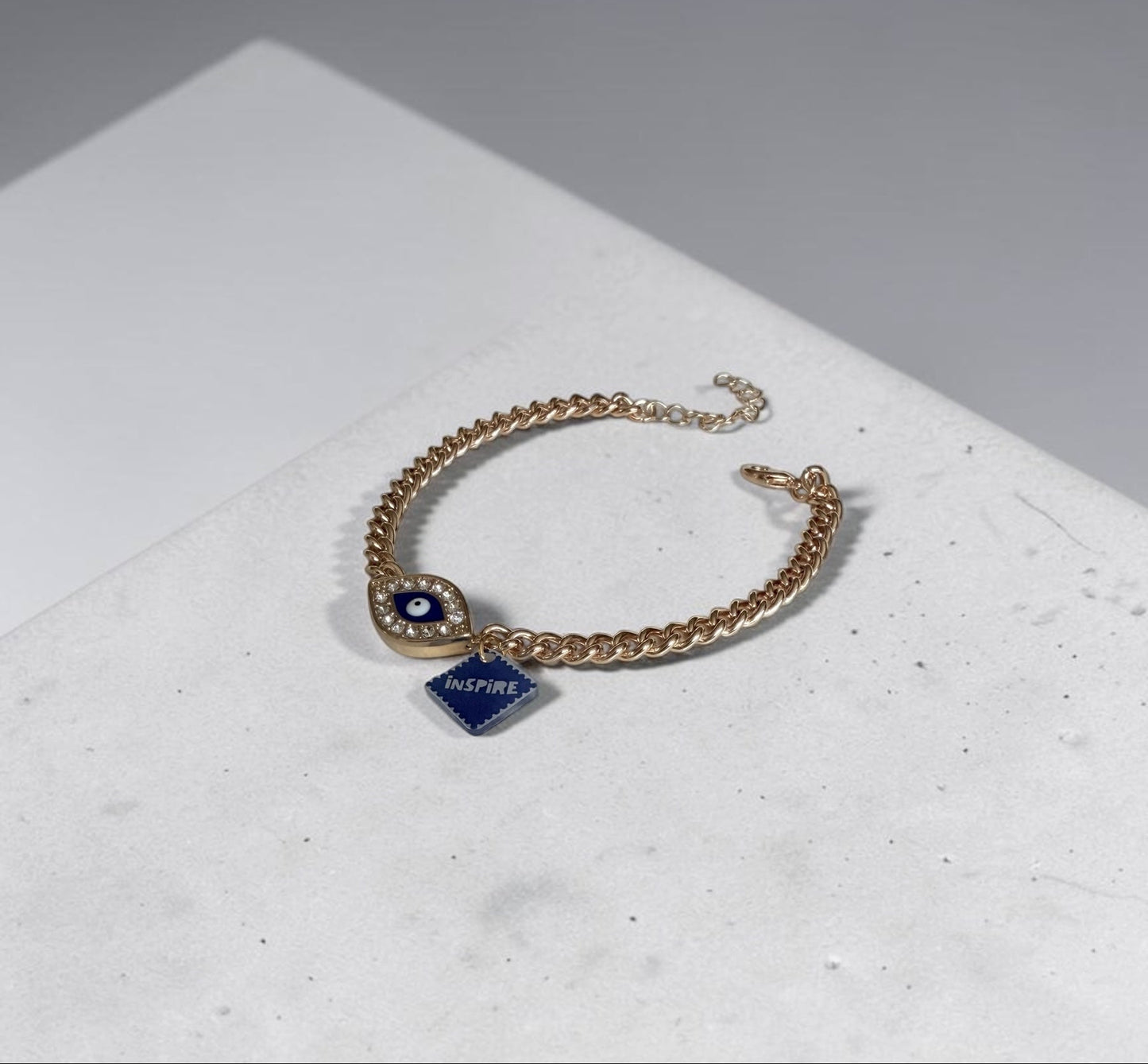 A gold chain anklet with a circular evil eye charm and a small blue diamond-shaped pendant engraved with 'INSPIRE', resting on a white textured surface.