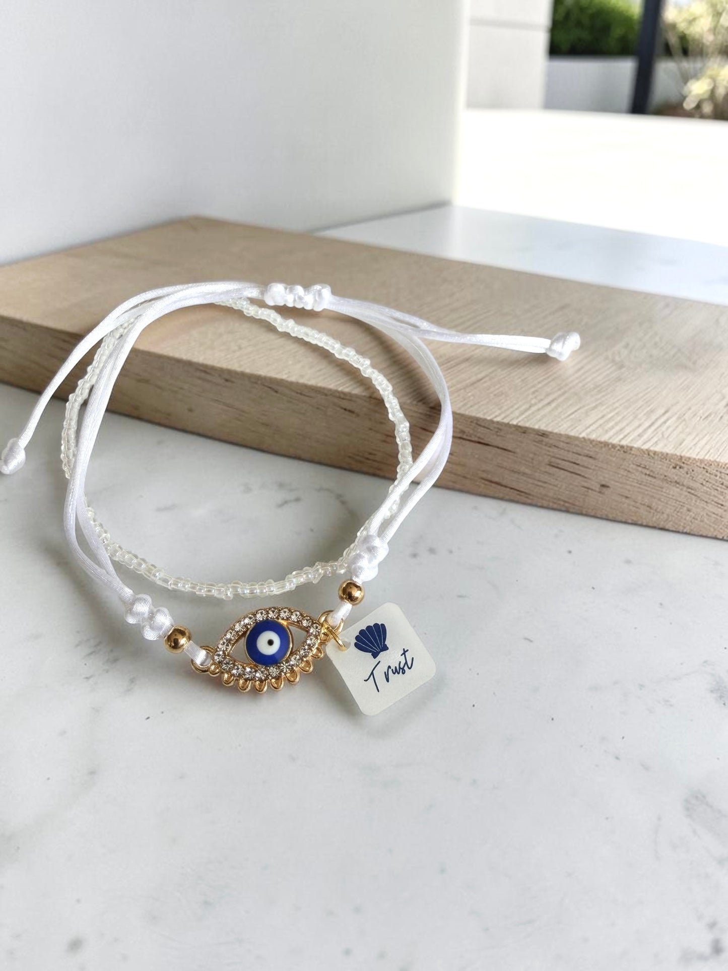 Two white bracelets on a wooden block: one with a cubic zirconia eye charm and 'Trust' tag, the other a plain seed bead design.