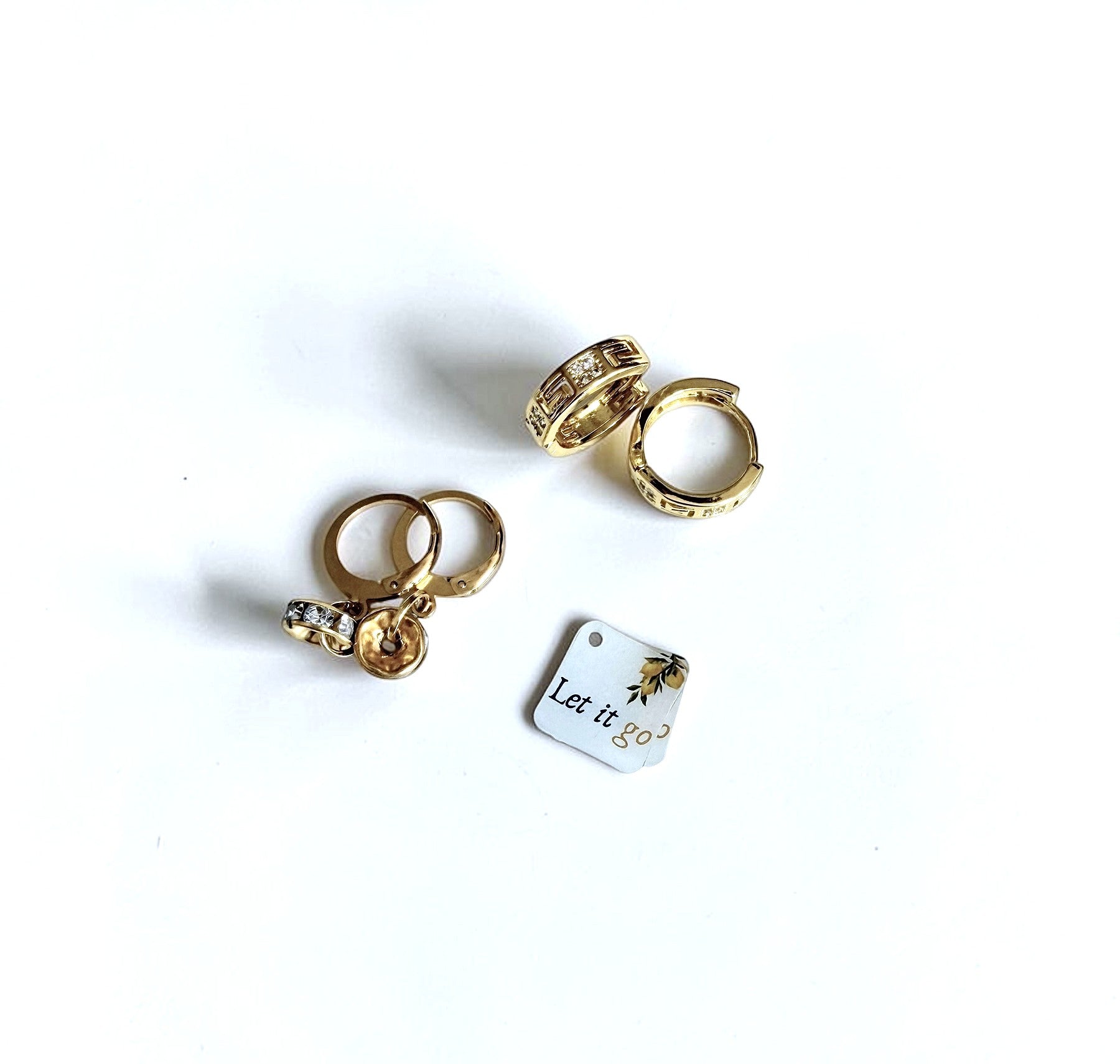 Two pairs of gold hoop earrings with textured and pave-set details, displayed alongside a small white tag that reads 'Let it go' with a lemon tree illustration, on a plain white background.