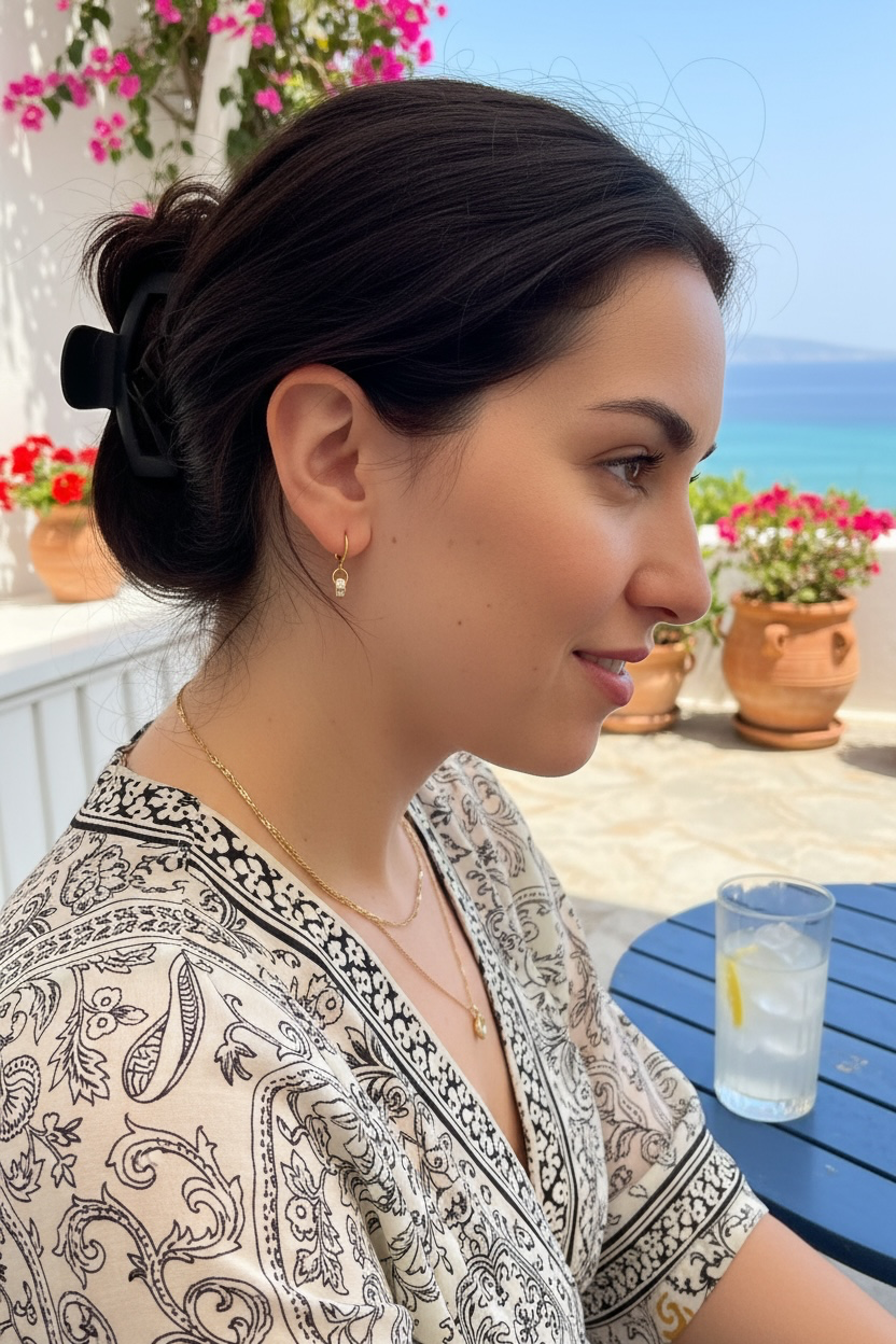 A woman in profile wearing a beige and black paisley patterned top, a simple gold earring with cubic zirconia set stones, and she is sitting at a blue table with a glass of water on a sunny outdoor patio with the sea in the background.