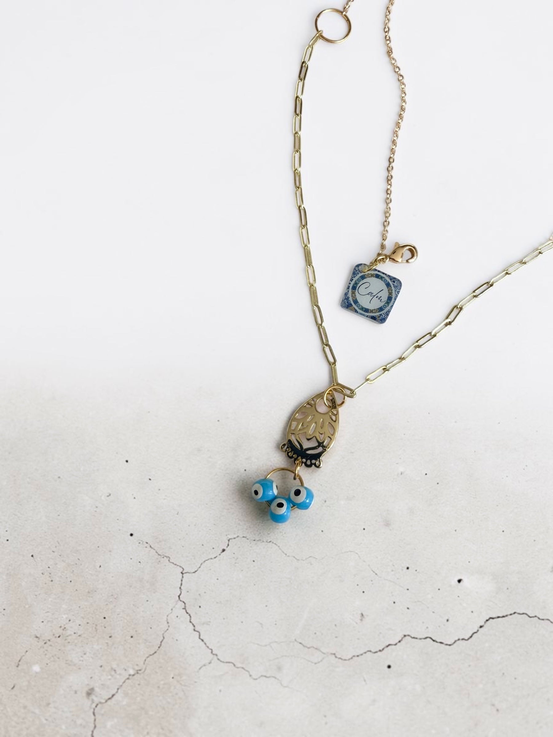 A delicate gold-toned necklace featuring an openwork gold geometric charm, a blue pendant labelled ' Calm', and blue evil eye beads, displayed on a light concrete surface.