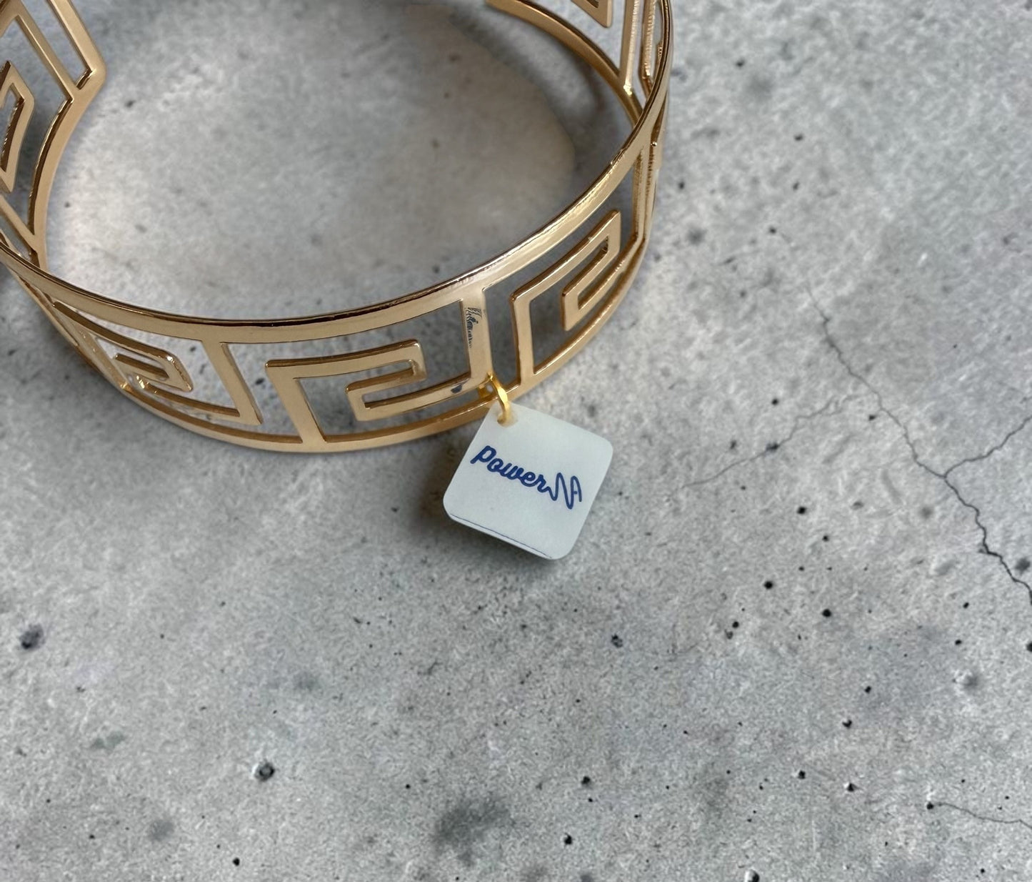 Aerial view of a gold Greek key bangle on a marbled grey surface, featuring a white tag labelled 'Power'.
