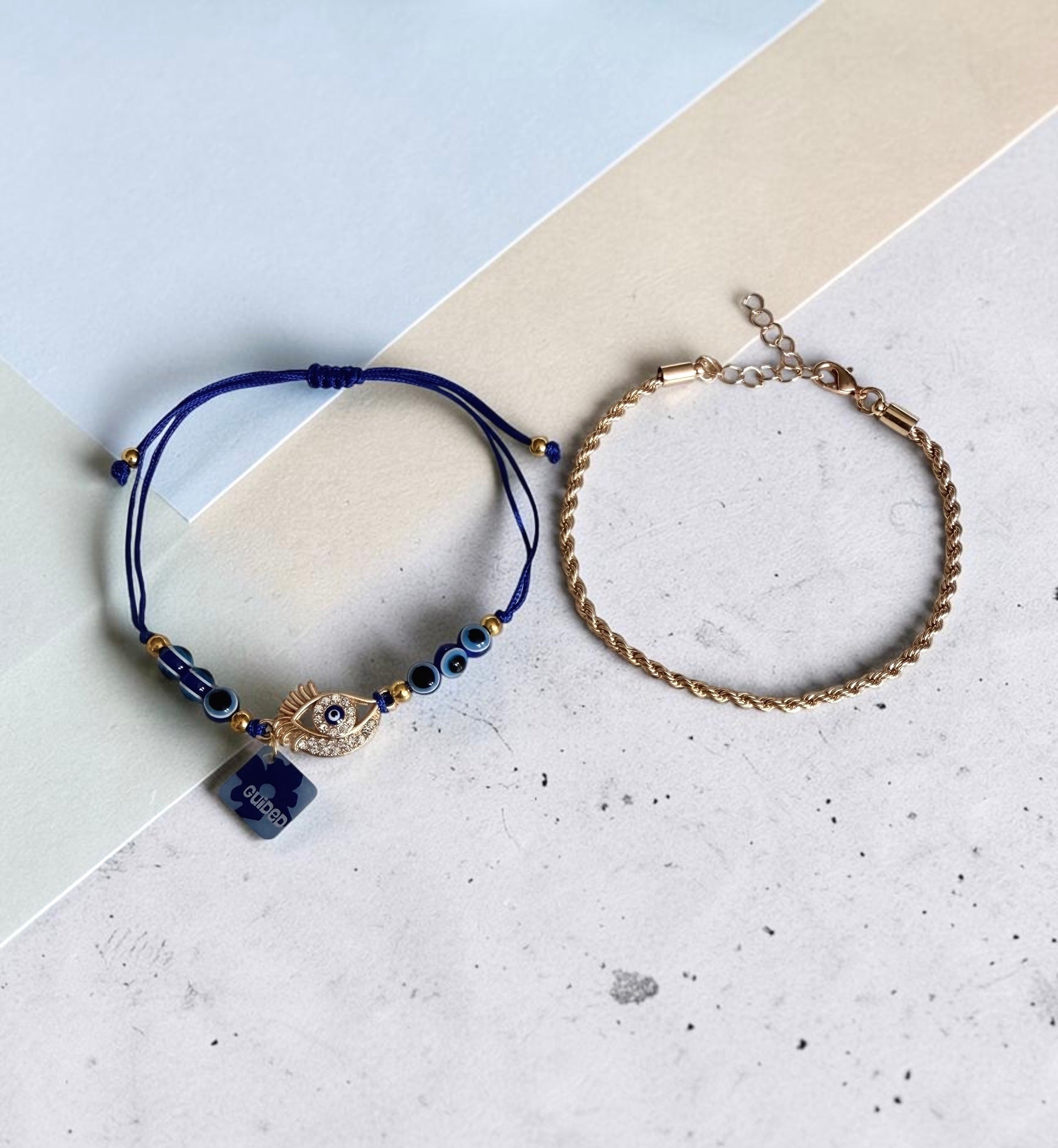 Two delicate anklets displayed on a split cream and light blue background. One anklet is dark blue with a central gold evil eye charm and a small blue stone pendant. And the other anklet is a plain gold twisted rope chain.