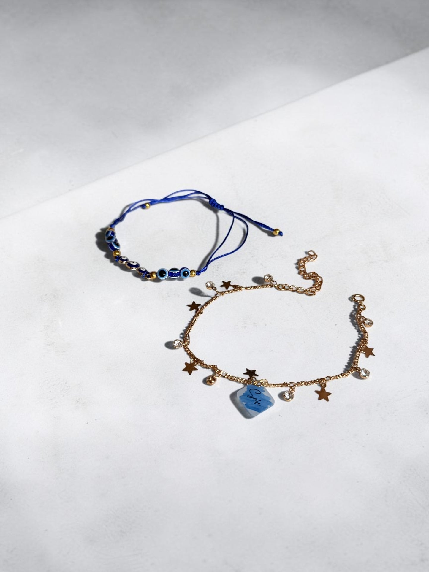 Two delicate jewellery pieces on a light grey surface: a blue cord anklet with evil eye beads and a gold-toned anklet with star charms, a small blue stone, and clear beads.